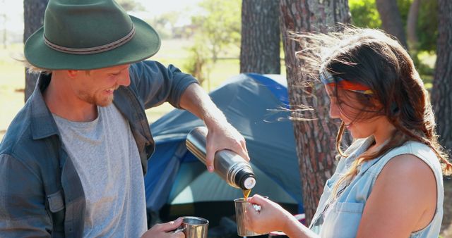 Couple enjoying hot drinks while camping outdoors during daytime. Man pouring beverage from thermos into mugs, with trees and a tent in the background. Suitable for travel blogs, camping advertisements, outdoor activity promotions, and lifestyle articles.