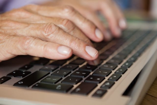 Close-up of senior woman's hands typing on a laptop keyboard. Ideal for illustrating themes related to elderly people using technology, digital literacy among seniors, remote work, online communication, and modern lifestyle for older adults.