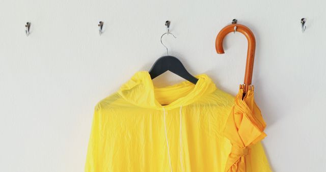 The image shows a yellow raincoat and a yellow umbrella with a wooden handle hanging on a wall with hooks. It is perfect for illustrating topics related to rainy weather, outdoor gear, and preparedness. Ideal for use in articles, blog posts, advertisements, and websites focused on weather, attire, and seasonal clothing.