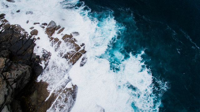 Aerial view showcasing powerful waves crashing against a rugged, rocky shoreline with vibrant blue ocean water and frothy white foam. Perfect for websites and blogs about nature, travel, oceanography, and coastal environments. Ideal for use in promotional materials or as a background image emphasizing natural beauty and dramatic seascapes.