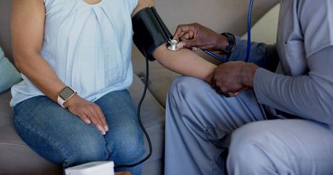 Home health clinician measuring blood pressure for adult patient on sofa with stethoscope