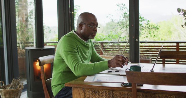 African american senior man sitting at dining table working, using laptop and drinking coffee. retirement lifestyle, spending time alone at home.