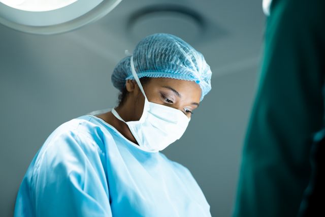 This image depicts an African American female surgeon wearing a face mask and surgical gown, deeply focused during a surgical procedure in an operating theatre. It can be used in healthcare-related content, medical service promotions, hospital websites, and educational materials about surgery and medical professions.