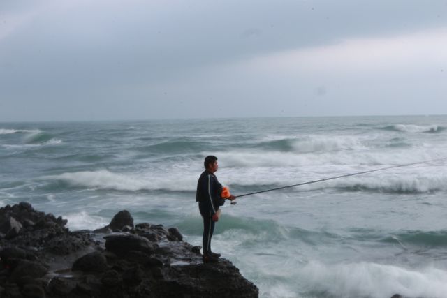 Man stands on rocky shoreline fishing with ocean waves crashing nearby during an overcast day. Ideal for concepts related to outdoor activities, fishing hobbies, relaxation, connection with nature, and coastal lifestyles.