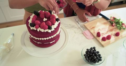 Diverse women decorating three-tier red velvet layer cake with fresh berries, baking joy