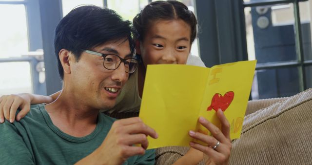 Father and daughter reading a colorful handmade greeting card at home, likely celebrating a special occasion. Suitable for themes of family bonding, parenting, loving relationships, and special moments in life. Great for use in advertisements, social media posts, articles on fatherhood, and lifestyle blogs focusing on family and parenting.