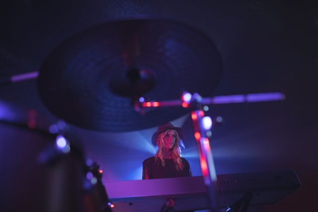 Female musician performing on stage in a dimly lit nightclub, illuminated by colorful lights. She is playing a keyboard and wearing a hat, creating an artistic and vibrant atmosphere. Ideal for use in articles, blogs, or promotional materials related to live music, nightlife, entertainment, and music events.