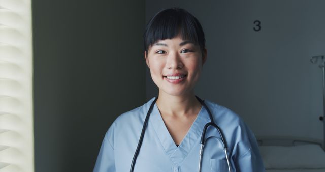 Asian female nurse wearing blue scrubs smiles with confidence in hospital. Stethoscope hanging around neck, highlighting professional medical role. Ideal for healthcare advertisements, hospital websites, and medical-related marketing materials showcasing dedicated healthcare professionals.