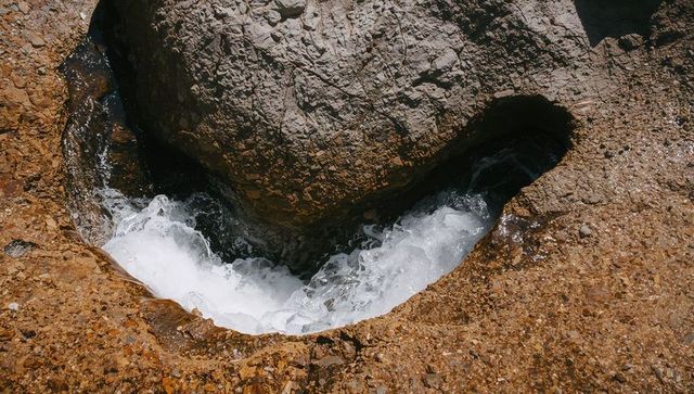 Sunlit coastal pothole with foaming water flowing through rounded rock cavity and pebbles
