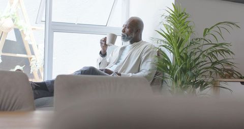 Senior african man enjoying morning coffee on sofa beside sunlit window with indoor plant