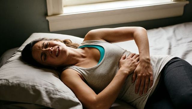 Young woman clutching abdomen on bed grimacing from menstrual cramp and abdominal pain