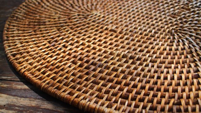 This close-up view highlights the intricate texture of a woven rattan placemat resting on a wooden table. Suitable for themes involving home decor, craftsmanship, natural materials, and rustic aesthetics. Ideal for blogs, design inspiration, furniture catalogs, and DIY project resources.