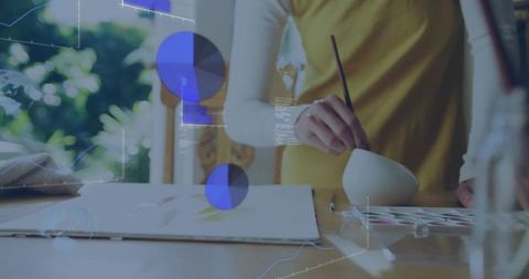 Artist dipping brush into bowl with watercolor palette and digital data overlays in studio
