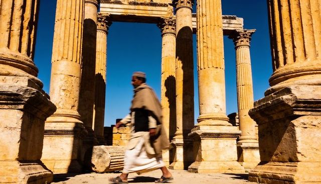 Walking man in traditional robe passing sunlit sandstone colonnade at ancient ruins