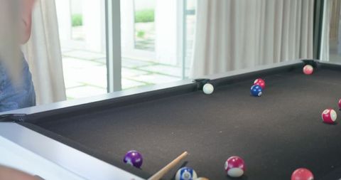 Woman in jeans leaning over black-felt pool table lining up cue shot in sunlit home