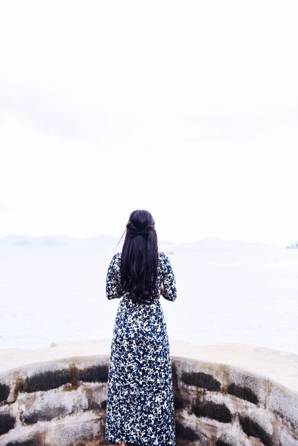 Woman with long hair in a floral dress standing by a stone barrier, looking out at the ocean. Suitable for themes of introspection, travel, seaside locations, fashion, and solitude.