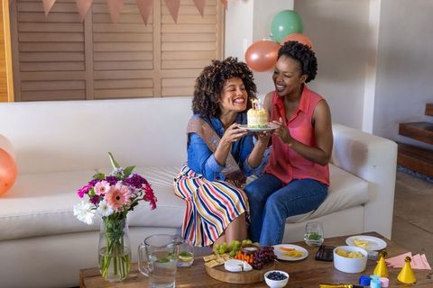 Black friends celebrating birthday at home holding lit cake with balloons