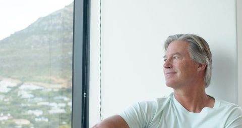 Senior man gazing through window at hillside view, peaceful reflective home moment