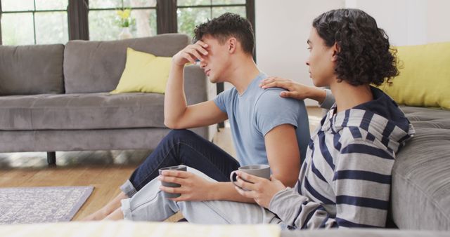 Worried diverse male couple sitting on floor and drinking coffee in living room. Spending quality time at home.