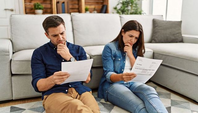 Young couple sitting on living room floor reviewing bills, calculating finances, worried