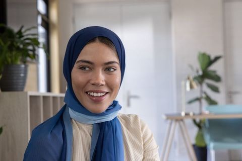 Confident muslim woman in blue hijab smiling in modern office workspace with greenery
