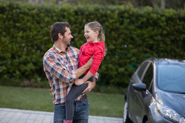 Father carrying daughter in his arms, both smiling and enjoying time together. Ideal for use in family-oriented advertisements, parenting blogs, and lifestyle articles. The casual setting with a car and greenery in the background adds a relatable and warm touch.