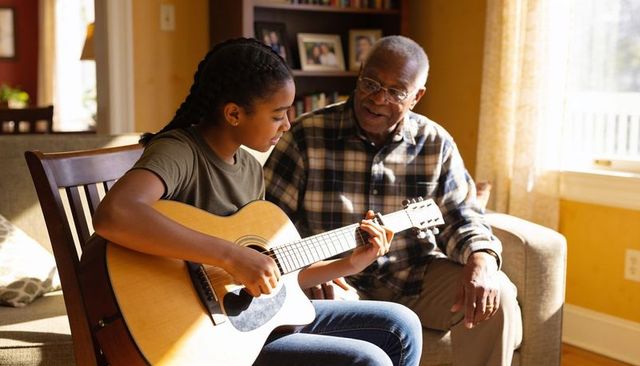 Grandfather teaching teen granddaughter guitar in sunny living room