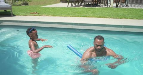 African father and son playing in backyard pool using foam noodle for joyful bonding