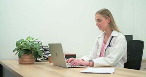 Female clinician typing on laptop at minimalist clinic desk wearing coat and stethoscope