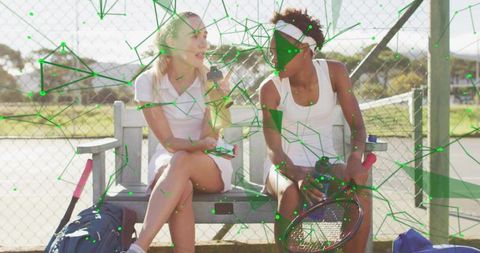 Courtside conversation: women talking on bench in white tennis outfits holding rackets and bottles