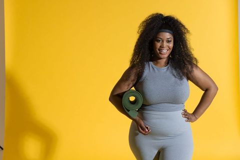 Smiling african american woman posing with green yoga mat in grey activewear on yellow backdrop