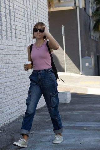 Young woman walking with coffee and backpack urban sidewalk wearing pink top and cargo jeans
