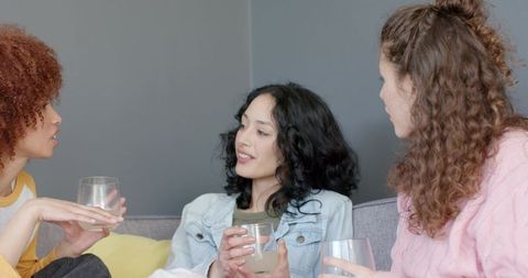 Diverse friends chatting and sipping drinks on gray sofa enjoying casual weekend gathering