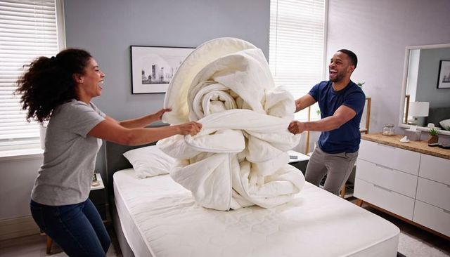 Couple laughing pulling giant knotted duvet on bed in bright modern bedroom