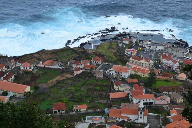 This vivid photo captures a scenic aerial view of a coastal town characterized by traditional red-roofed houses and a rugged, picturesque shoreline with crashing ocean waves. Ideal for illustrating travel guides, tourism content, countryside architecture, and coastal landscapes. Perfect as a visual element for blogs, magazines, and websites focusing on European travel and scenic locations.