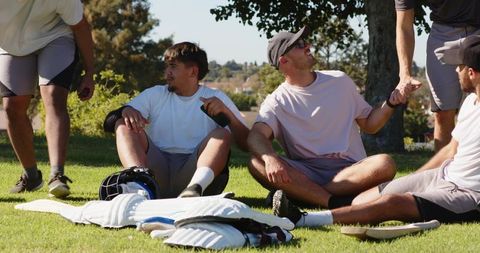Cricket teammates sitting on grass under tree chatting, relaxing with gear and camaraderie