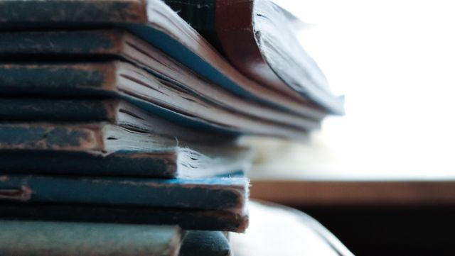 Stack of old, worn books lying on a wooden desk in soft focus. This nostalgic and serene composition evoking a sense of timelessness is perfect for illustrating themes relating to education, literature, or lifelong learning. It can be used in articles, blog posts, advertisements, and prints celebrating knowledge, libraries, reading habits, and vintage aesthetics.