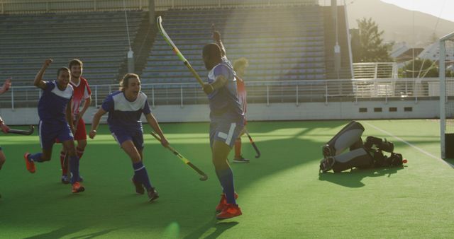 Joyful field hockey players in blue uniforms celebrating a victorious moment on green turf with excitement. The sun lit stadium amplifies their joy and teamwork spirit. Ideal for sports motivation themes, team dynamics discussions, and victory-based visuals for marketing material.