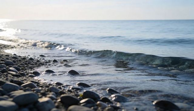 Pebble shoreline washing by gentle wave with sunlight glinting on calm blue sea