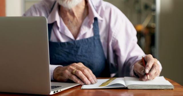 A senior Caucasian man in an apron writes in a notebook beside a laptop, with copy space. His focus and attire suggest he could be a craftsman or artist planning his work or managing orders.