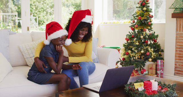 Mother and daughter wearing Santa hats sitting on sofa looking at laptop, festive Christmas tree and decorations in background. Perfect for highlighting family holiday moments, virtual celebrations, and the joy of togetherness during the festive season. Suitable for Christmas cards, holiday advertisements, and online greetings.