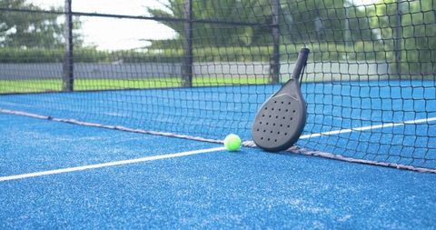 Perforated padel paddle leaning against net with neon ball on blue outdoor court