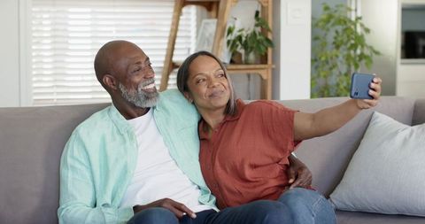 Mature black couple taking selfie on sofa in sunlit living room, cozy home lifestyle moment