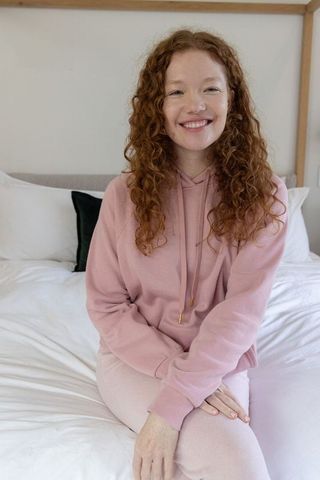 Red curly-haired woman smiling on bed in pink hoodie cozy minimalist bedroom lifestyle