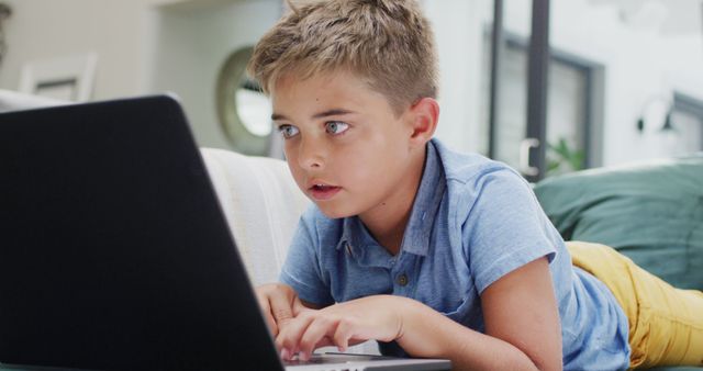 Young boy lying on a couch, using a laptop, with a focused expression on his face. This image can be used for educational materials, childhood technology use, online learning platforms, or articles discussing children's screen time or digital entertainment.