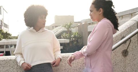 Two women chatting on urban overpass railing backlit in cream top and pink hoodie
