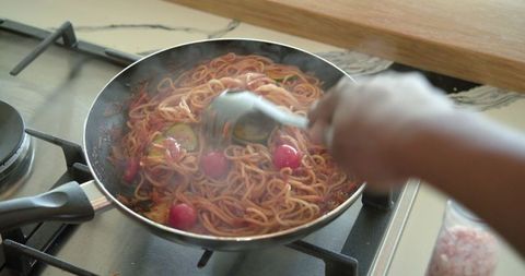 African american woman stirring tomato spaghetti with zucchini in skillet on gas stove
