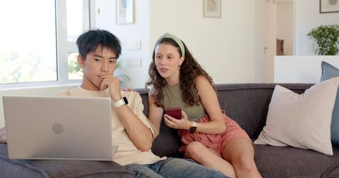 Young couple sitting on couch browsing laptop and smartphone, planning together at home