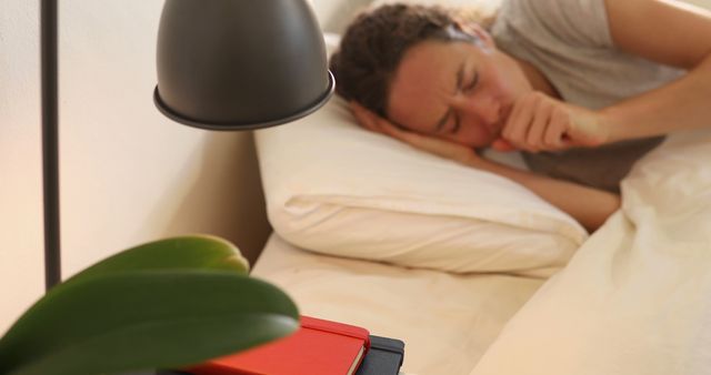 A woman is lying in bed, wrapped in a cozy white blanket, and is coughing. The bedside table next to her holds a lamp and red notebooks. This image can be used for themes related to health, illness, relaxation, and home environments. Ideal for articles or advertisements about recovery, healthcare products, and wellness tips.