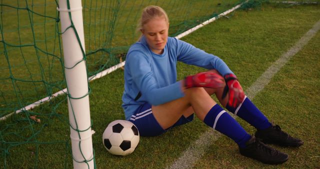 A young female soccer goalkeeper is relaxing on the grass near the goal after a practice session. She has her gloves on and is sitting next to a soccer ball. This image can be used for sports-related promotions, youth sports campaigns, athletic training materials, or articles on women in sports.